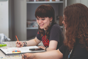 Two students sat at a desk together smiling and doing artwork