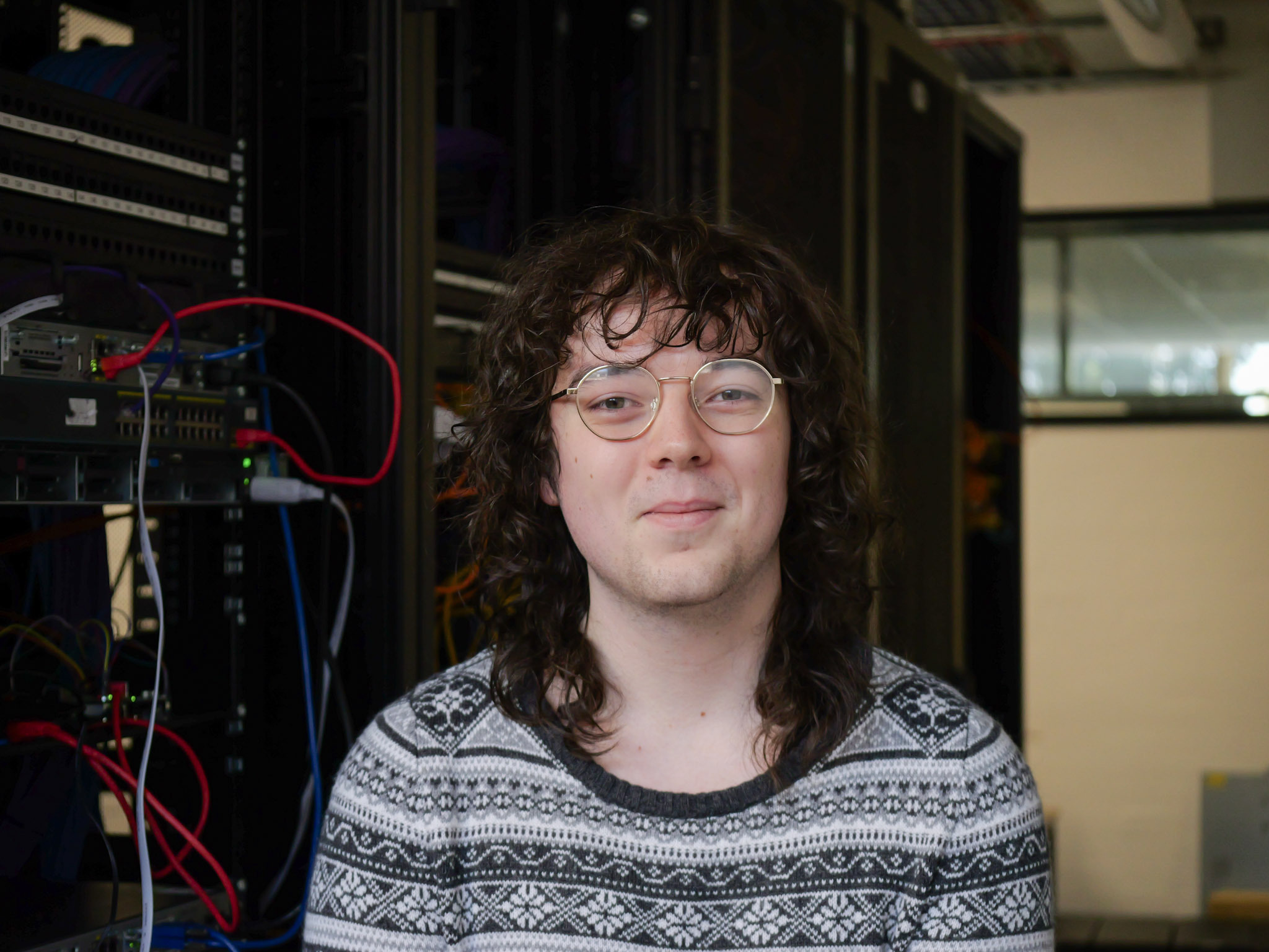A person with brown curly hair and wire rimmed glasses smiles in front of a server room.