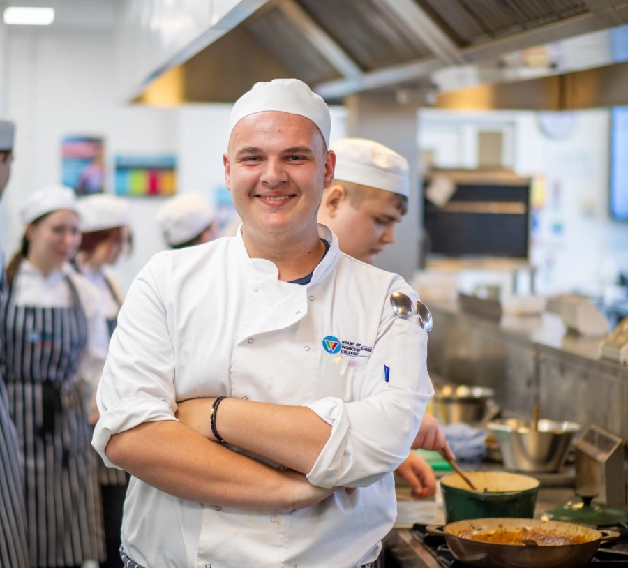 Student smiling in chefs uniform in kitchen with other students blurred in background