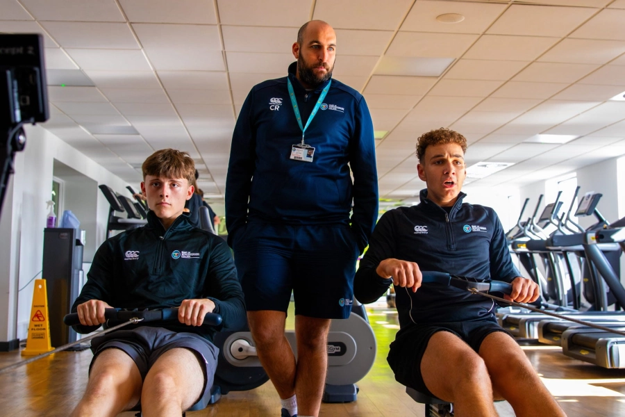 Two students on rowing machines with teacher watching from behind