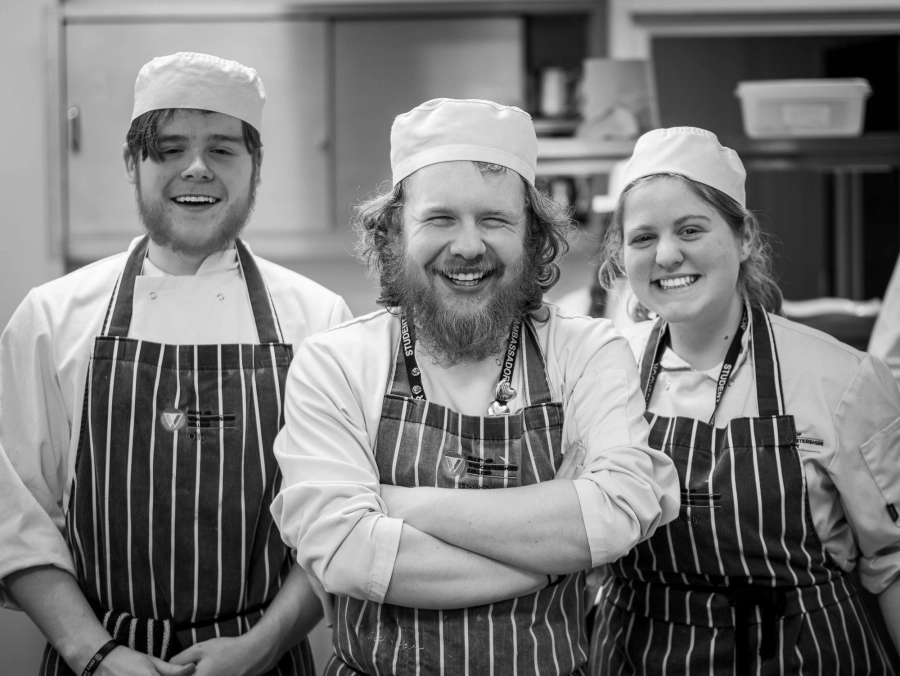 3 student chefs posing in chefs uniform in a kitchen