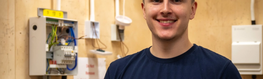 Picture of electrical installation student stood in electrical workshop with wirings behind