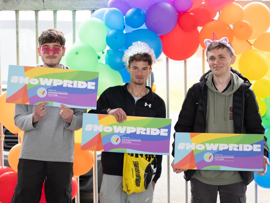 3 students at a pide event at college holding up #HoWPride boards wearing sunglasses and headbands