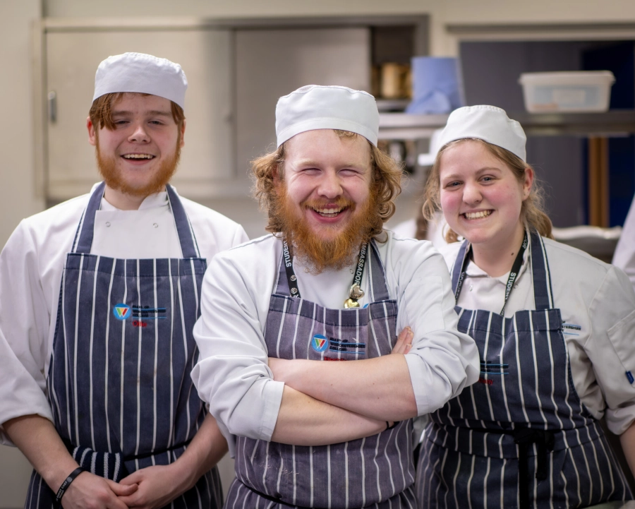 Three students in their hospitality and catering uniforms smiling in the kitchens for a picture.