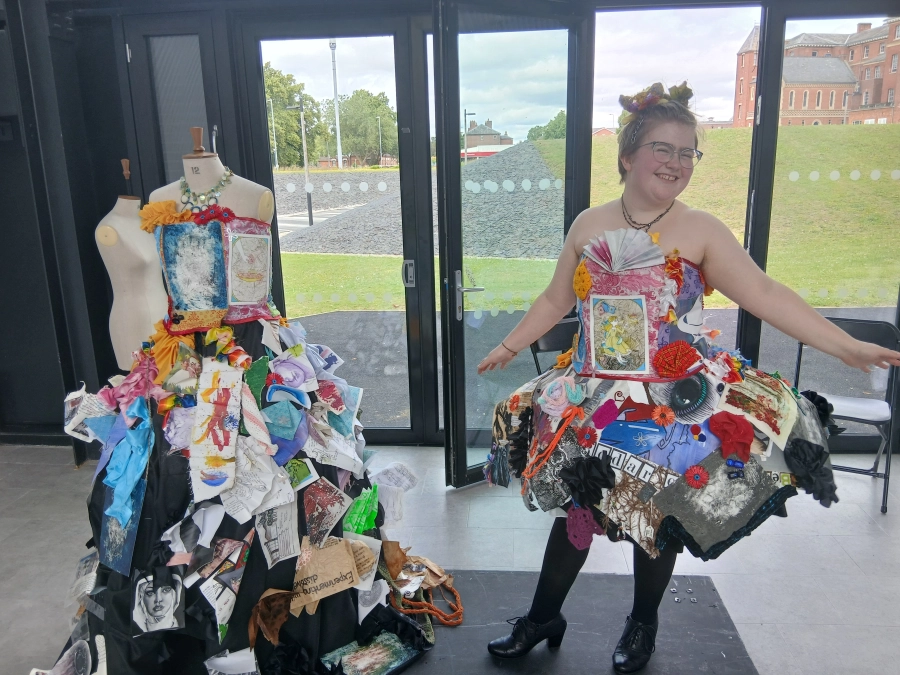 An art and design student stood next to a dress made of paper images and also wearing a similar dress made from paper images.